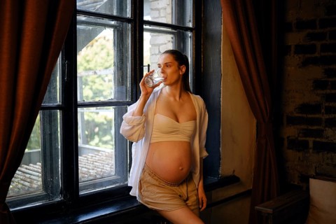 A pregnant woman stands by a large window at home, enjoying a drink from a glass. She wears a