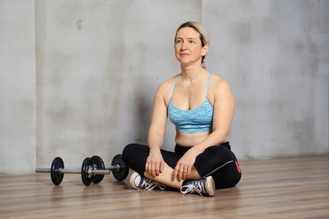 A woman sits cross-legged on a wooden gym floor, dressed in workout attire. Nearby, a pair of
