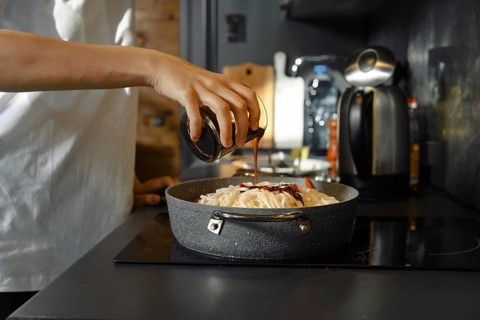 Unrecognizable person adds a soy sauce to a skillet filled with fresh vegetables and noodles on the