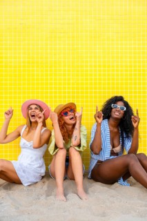 Three diverse women wearing summer clothes and sunglasses are sitting on the sand, pointing upwards