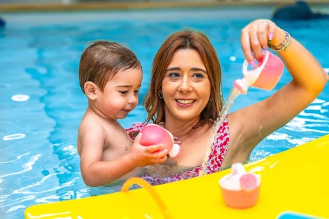 Happy mother and son having fun, playing with pink toys and pouring water in a swimming pool during