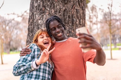 Two happy young students are making the victory sign while taking a selfie with a smartphone in a