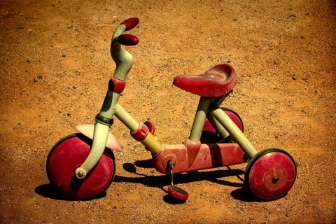 A vintage red tricycle sits abandoned on a sandy ground, showing signs of wear and tear, evoking