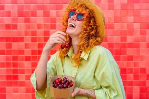 Young redhead woman with heart shaped sunglasses and straw hat is laughing while eating fresh