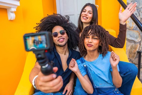 Three happy multi ethnic influencers sitting on yellow steps are filming a vlog with an action