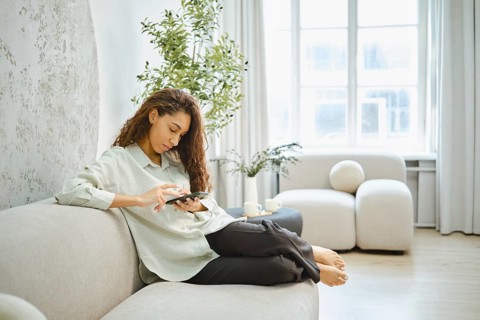 In a bright living room, a woman with curly hair sits comfortably on a light sofa, focused on her
