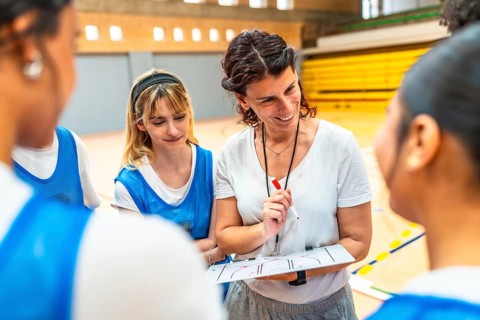 Smiling female coach explaining basketball game strategy to her team using a clipboard in a gym
