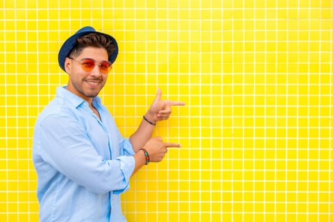 Fashionable man wearing a hat and sunglasses points to the side against a vibrant yellow tiled