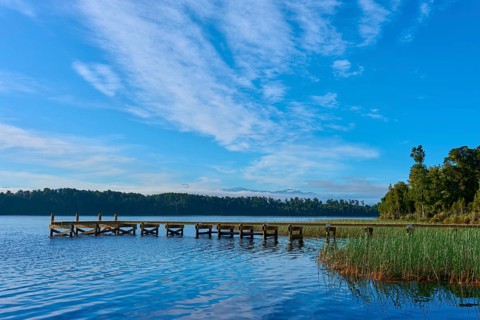 A jetty leads into a calm lake under a blue sky with light clouds, surrounded by lush nature, Lake