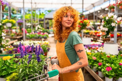 Young woman pushing a shopping cart while selecting vibrant flowers in a garden center greenhouse,