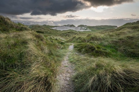 A narrow sandy path leads through a grassy landscape with dunes and dark clouds, near Hvide Sande,