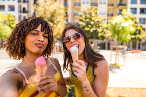 Two cheerful women enjoying ice cream cones outdoors, taking a selfie on a sunny summer day in an