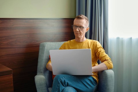 Middle-aged man in a yellow shirt sits comfortably in a grey armchair, focused on his laptop.