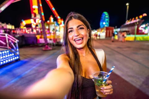 Happy young woman taking a selfie while enjoying a refreshing drink at a vibrant amusement park at