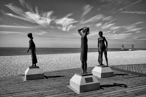 Bronze sculpture La Heure du Bain by Dominique Denry on the beach, sky, blue, behind it beacon Feu