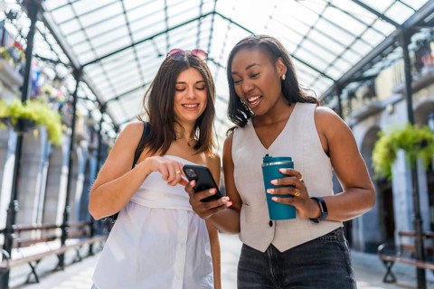 Two happy businesswomen walking through a city gallery, sipping coffee and using a smartphone,