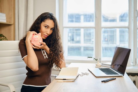 Cute woman with long curly hair is smiling and holding a pink piggy bank. She is seated at a