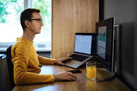 A man wearing glasses sits at a wooden desk in a home office, intently working on a laptop and