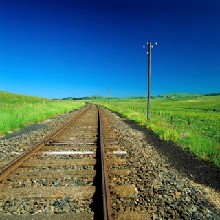 The railway track stretches through a lush green rural area, flanked by vibrant grasslands and a