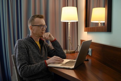 Middle-aged man sits at a wooden desk in a hotel room, focused on his laptop and is watching a