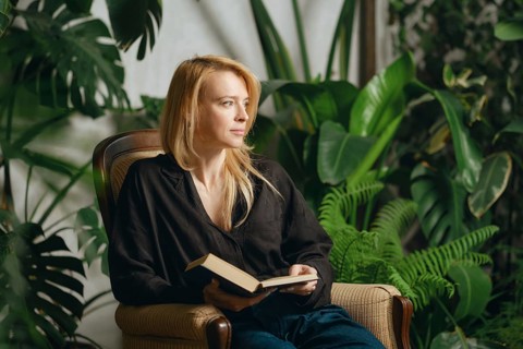 A woman sits comfortably in an armchair holding a book, surrounded by vibrant green plants.She is