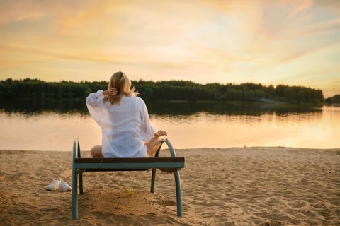 A woman sits peacefully on a bench near the riverbank, enjoying the serene sunset. The soft golden