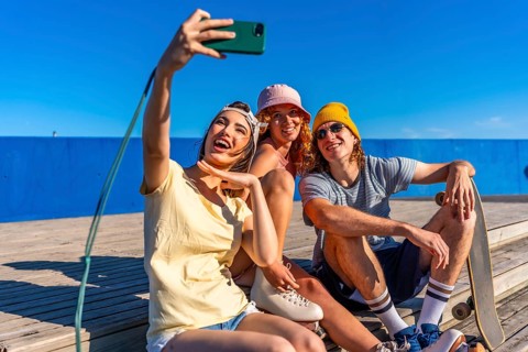 Three cheerful young friends are sitting on a wooden walkway by the sea, taking a selfie with a