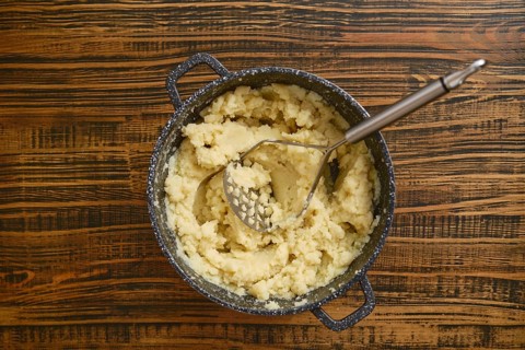 Creamy mashed potatoes are freshly prepared in a pot, sitting on a rustic wooden table. A potato