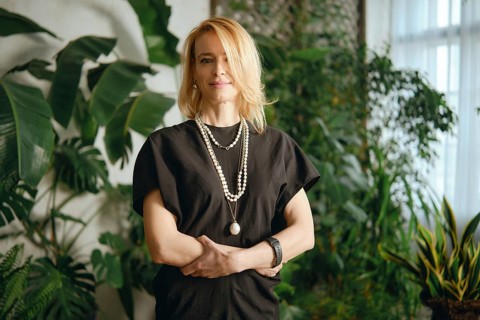 A woman stands gracefully amidst lush greenery in a cozy indoor garden. Her stylish outfit is