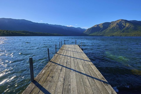 A narrow footbridge leads into the blue waters of a lake, surrounded by an impressive mountain
