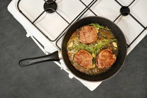 Three thick beef steaks are searing in a skillet on a gas stove. Fresh herbs and garlic surround