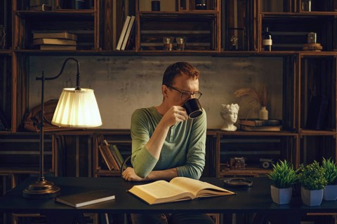 A middle-aged man sits at a desk in a cozy room, sipping a herbal tea from a mug. Soft light from a