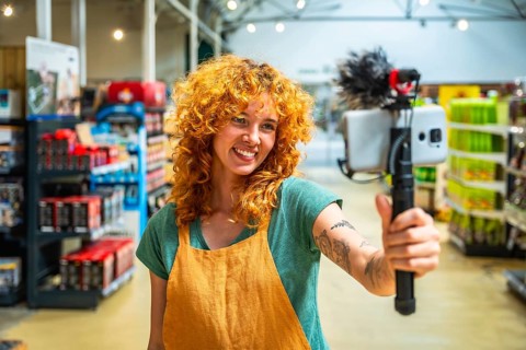 Young woman working as supermarket clerk vlogging using smartphone and microphone promoting