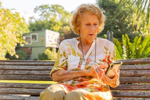 Senior woman sitting on a wooden bench in a sunny park, using a mobile phone to stay connected