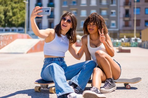 Two young women are taking a selfie with a smartphone while sitting on their skateboards in a skate