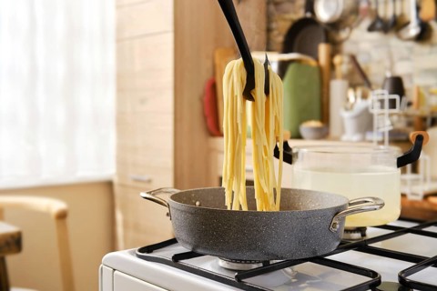 A person lifts cooked spaghetti from a pot using a utensil, with steam rising in a cozy kitchen.