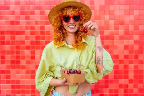 Happy young woman with curly red hair eating cherries from a small cardboard box, wearing heart