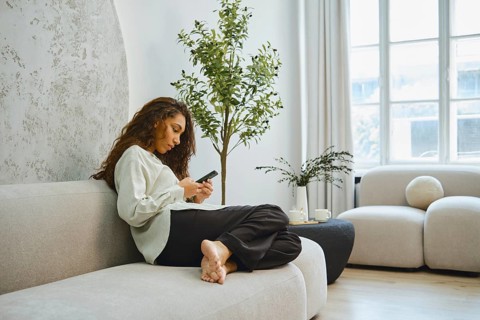 A woman sits comfortably on a light-colored sofa, engaged with her smartphone. She is in a spacious