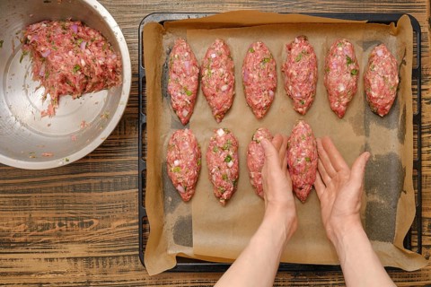 Freshly shaped kebabs are arranged on a baking tray lined with parchment paper. A mixing bowl