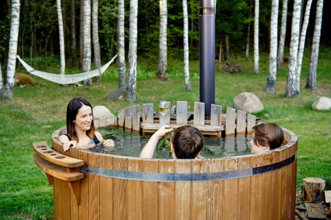 Two children and an their mom enjoy a warm soak in an outdoor wooden hot tub. Lush green grass and