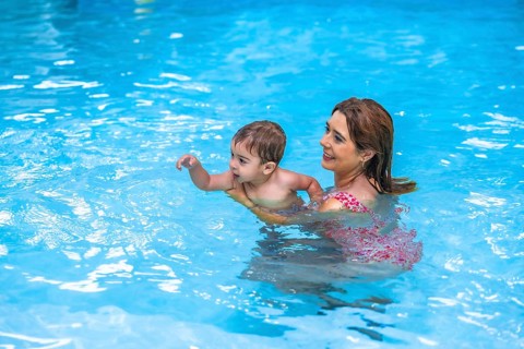 Happy mother holding her toddler son while teaching him to swim in a pool with clear blue water