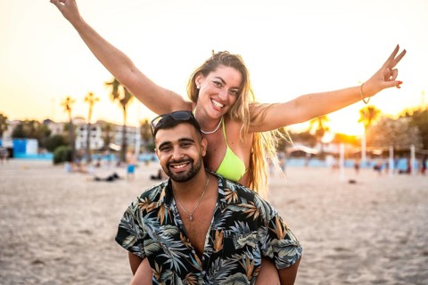 Young couple enjoying piggyback ride on beach, woman making victory sign, sunset in background