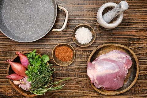 Fresh turkey thigh on a wooden plate alongside vibrant herbs, shallots, and spices. Overhead view