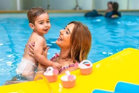Mother holding her smiling son in a swimming pool next to floating toys, enjoying a playful moment