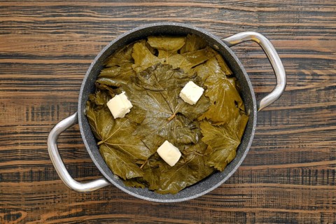 A pot filled with grape leaves prepares to cook dolma on a wooden table. Butter sits on top, ready