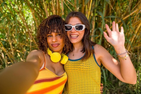 Two happy young women with headphones and sunglasses are taking a selfie and waving in a bamboo