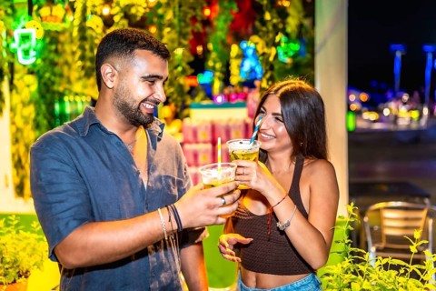 Young man and woman smiling and toasting with cocktails in a vibrant night market setting,