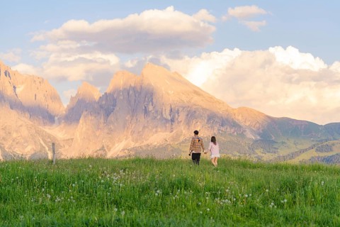 A couple walks across a flowering meadow towards the mountain range at sunset, Alpe di Siusi,
