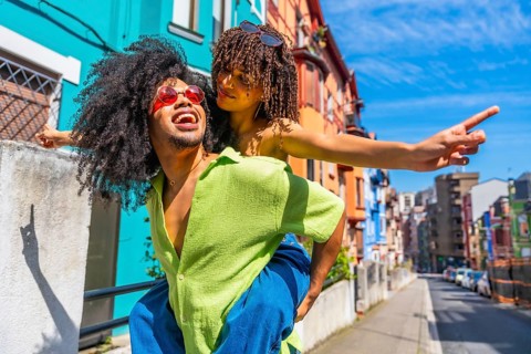 Happy tourists having fun piggybacking in a colorful city street, enjoying summer holidays and