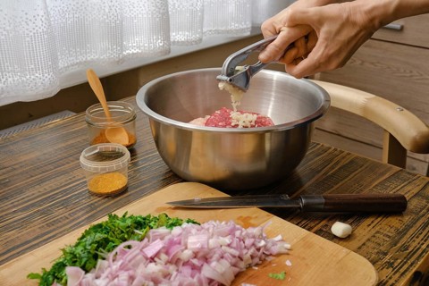 Hands mincing garlic into a metal bowl filled with ground meat while chopped onions and fresh herbs